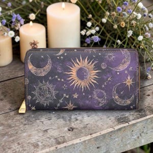 May include: A purple and black celestial-themed wallet featuring gold suns, moons, and stars. The wallet has a zipper closure with a gold pull and is displayed on a wooden surface. The background includes lit candles and floral arrangements.