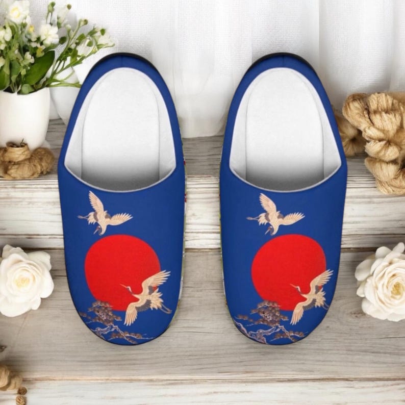 May include: A pair of blue slippers with a Japanese-inspired design. Each slipper features a red sun, white cranes, and a detailed tree branch design. The interior is white, and the slippers are designed for indoor use.