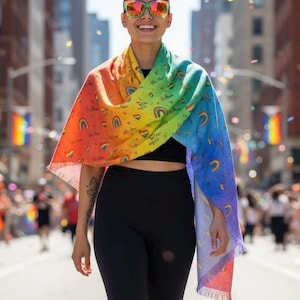 Könnte beinhalten: Ein regenbogenfarbener Schal mit Fransen, verziert mit Regenbogen- und Herzmotiven, getragen über einer schwarzen Leggings und einem schwarzen Oberteil. Die Person trägt eine Sonnenbrille mit farbigem Rahmen. Im Hintergrund eine Stadtstraße.