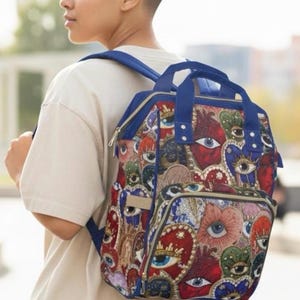 Może przedstawiać: Niebieski plecak z powtarzającym się wzorem oczu i serc w kolorach czerwonym, niebieskim, zielonym i złotym. Plecak ma niebieskie paski i górny uchwyt. Wzór jest przyciągający wzrok i kolorowy.