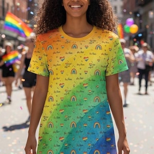 Op de afbeelding: Een regenboogkleurige t-shirtjurk met harten, regenbogen en de voornaamwoorden "she/her". De jurk, gedragen door een persoon met een zonnebril, loopt over van geel naar blauw. Op de achtergrond een Pride parade.