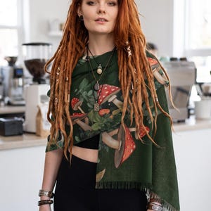 May include: A dark green rectangular scarf featuring a detailed print of red and white spotted mushrooms and green foliage. The scarf has a soft, woven texture with a fringed hem along the bottom edge, draped over a black top and leggings.