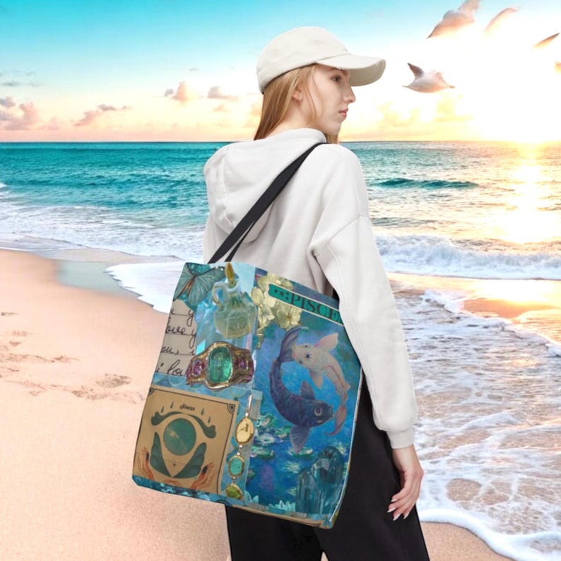 May include: A tote bag with a blue and teal design featuring a Pisces theme. The bag has a black shoulder strap and a variety of images, including a ring, a butterfly, and two fish. The bag is being carried on a beach.