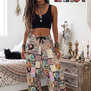 May include: Wide-leg pants featuring a colorful patchwork print of tarot card illustrations including suns, moons, and celestial motifs. The pants have a black drawstring waistband and are paired with a solid black sleeveless crop top.