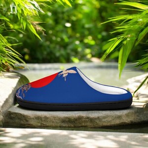 May include: A blue slipper with a red accent and a crane design. The slipper has a black sole and a white interior. The design includes a red circle and a crane in flight, set against a blue background.
