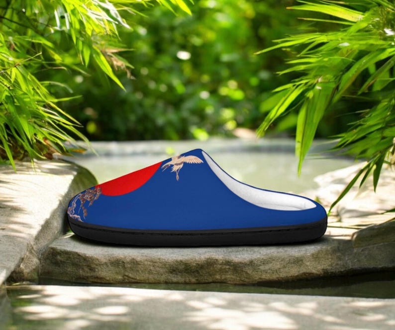 May include: A blue slipper with a red accent and a crane design. The slipper has a black sole and a white interior. The design includes a red circle and a crane in flight, set against a blue background.
