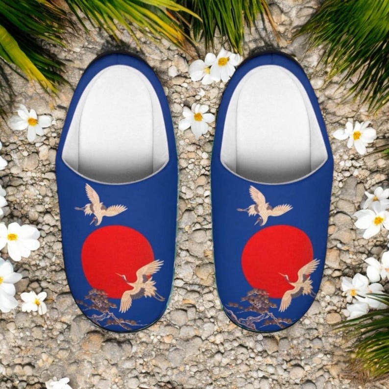 May include: A pair of blue slippers with a white interior. Each slipper features a red sun design with flying cranes and a detailed branch illustration. The slippers are surrounded by white flowers and greenery.