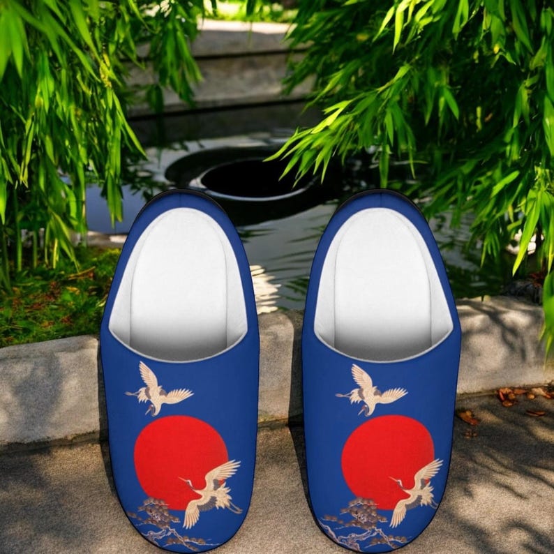 May include: A pair of blue slippers with a Japanese-inspired design. Each slipper features a large red sun, white cranes, and a detailed wave pattern. The interior is white, and the slippers are set against a natural outdoor background.