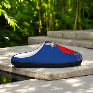 May include: A blue slipper with a white interior and a black sole. The slipper features a design of a red sun, a crane, and a landscape. The slipper is resting on a stone surface.