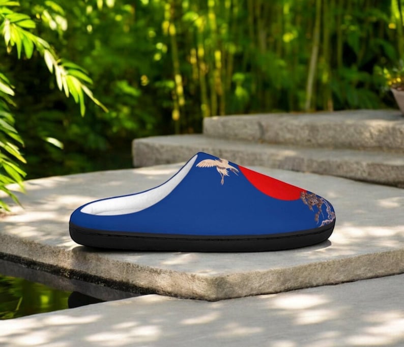 May include: A blue slipper with a white interior and a black sole. The slipper features a design of a red sun, a crane, and a landscape. The slipper is resting on a stone surface.