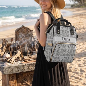 May include: A light grey backpack featuring a repeating black line art pattern of suns, birds, and mystical symbols. The bag has black straps, gold-toned zips, and a horizontal front pocket with the name Dana printed in black script lettering.