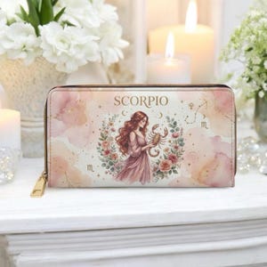 Pode incluir: Uma carteira rosa e dourada com zíper do signo de Escorpião. A carteira apresenta uma ilustração em aquarela de uma mulher segurando um escorpião, rodeada por flores. A palavra "SCORPIO" é impressa na parte superior.