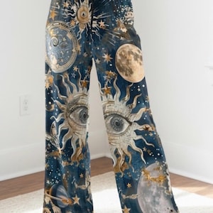 Op de afbeelding: Wijde loungebroek in diep marineblauw met een hemelse print. Het patroon bevat gouden zonnen met centrale ogen, verschillende maanfasen, verspreide sterren en planetaire ringen. De broek heeft een zwarte tailleband met trekkoord.