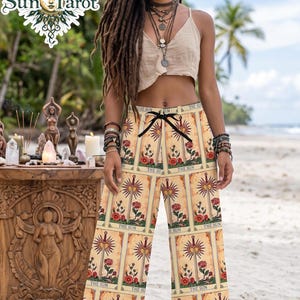 May include: Wide-leg pants featuring a repeating tarot card print with the text THE SUN, a central sword, and red roses on a light yellow background. The pants have a black drawstring waist and a loose, flowing fit, worn with a beige crop top.