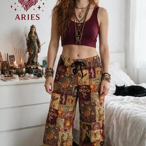 May include: A maroon sleeveless crop top paired with wide-leg pants featuring a multi-colored collage print of Aries zodiac symbols, ram illustrations, and text reading "ADMIT ONE" and "Aries". Matching patterned slippers are worn on the feet.