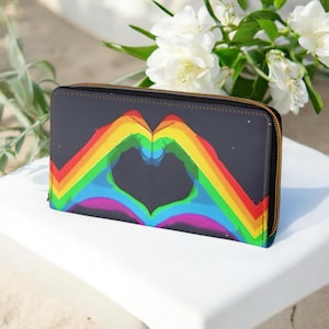 Puede incluir: Una cartera gris oscuro con un diseño de corazón arcoíris. La cartera tiene una cremallera dorada y una forma de corazón colorida hecha de colores del arcoíris. La cartera es rectangular y parece ser de cuero.