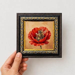 May include: A square framed painting featuring a detailed illustration of a bee sitting inside a vibrant red poppy flower whilst holding a small open book. The artwork is encased in a dark, ornate frame with gold-toned decorative accents.