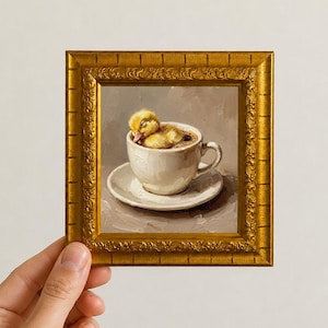 May include: An oil painting in a decorative gold frame depicting a small yellow duckling sleeping inside a white ceramic coffee cup on a matching saucer. The painting features visible brushstrokes and a neutral, muted background color palette.