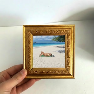 May include: A small, square, gold-framed painting depicts a person sunbathing on a white sand beach. The artwork showcases a turquoise ocean, blue sky, and a tree providing shade. The frame has ornate detailing.