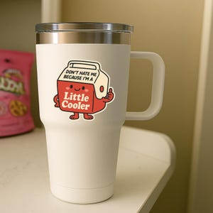 K&ouml;nnte beinhalten: Ein wei&szlig;er Isolierbecher mit silbernem Rand und Griff. Ein Aufkleber auf dem Becher zeigt einen Cartoon-K&uuml;hler mit dem Text "DON'T HATE ME BECAUSE I'M A Little Cooler". Der K&uuml;hler ist rot mit wei&szlig;en Akzenten und zeigt einen Daumen hoch.