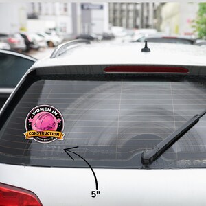 Puede incluir: Una pegatina redonda, de aproximadamente 12,7 cm de di&aacute;metro, est&aacute; adherida a la ventana de un coche. Muestra el texto "WOMEN IN CONSTRUCTION" alrededor de un casco rosa. Una pancarta debajo dice "STRONG & SKILLED".