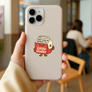K&ouml;nnte beinhalten: Beigefarbene Handyh&uuml;lle mit einem Cartoon-Aufkleber eines rot-wei&szlig;en K&uuml;hlers mit dem Text "Don't hate me because I'm a Little Cooler". Der K&uuml;hler zeigt einen Daumen hoch.
