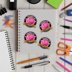 Puede incluir: Cuatro pegatinas redondas con un dise&ntilde;o de casco rosa, el texto "WOMEN IN CONSTRUCTION" y "STRONG & SKILLED". Las pegatinas est&aacute;n sobre un cuaderno gris, rodeadas de material de oficina sobre una superficie de madera.