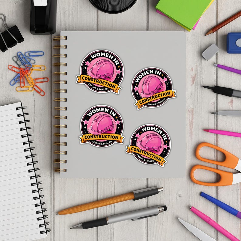 Puede incluir: Cuatro pegatinas redondas con un dise&ntilde;o de casco rosa, el texto "WOMEN IN CONSTRUCTION" y "STRONG & SKILLED". Las pegatinas est&aacute;n sobre un cuaderno gris, rodeadas de material de oficina sobre una superficie de madera.