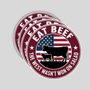 May include: A stack of round stickers with a burgundy border and white text that reads "EAT BEEF" and "THE WEST WASN'T WON ON SALAD". The design includes a silhouette of a cow and calf in front of an American flag.