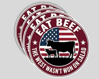 May include: A stack of round stickers with a burgundy border and white text that reads "EAT BEEF" and "THE WEST WASN'T WON ON SALAD". The design includes a silhouette of a cow and calf in front of an American flag.