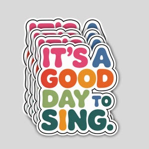 May include: A stack of colorful stickers with the phrase "IT'S A GOOD DAY TO SING" in bubble letters. The stickers have a white border and are in shades of pink, orange, green, blue, and teal.
