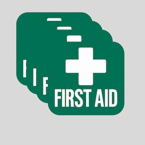 May include: A stack of green, square first aid signs with rounded corners. Each sign features a white cross and the words "FIRST AID" in white, centered below the cross. The background is a solid light gray.