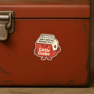 K&ouml;nnte beinhalten: Ein Aufkleber mit einem Cartoon-K&uuml;hler mit dem Text "Don't hate me because I'm a Little Cooler." Der K&uuml;hler ist rot mit einem wei&szlig;en Deckel und einem Daumen hoch. Der Aufkleber befindet sich auf einer rostig roten Oberfl&auml;che.