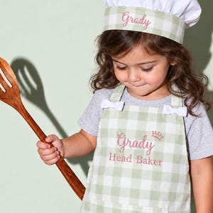 May include: A sage green and white gingham apron and matching chef hat set. The apron features a front pocket, two small white bows at the shoulders, and embroidery reading "Grady Head Baker" with a crown. The hat is embroidered with "Grady".