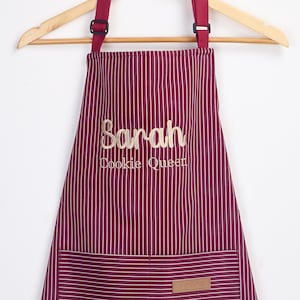 May include: A burgundy apron with thin white vertical stripes, featuring adjustable dark red neck straps and a front pocket. The text Sarah is embroidered in gold script above the gold embroidered text Cookie Queen. A small tan label is on the pocket.