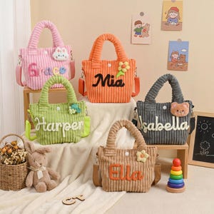 May include: Five corduroy tote bags in pink, orange, green, gray, and tan, each with a name embroidered on the front. The bags have matching handles and adjustable shoulder straps. Decorative accents include flowers and a bunny.
