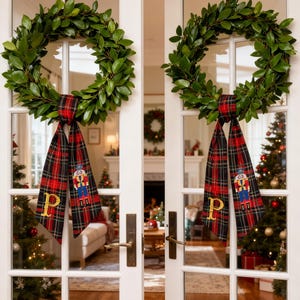 Puede incluir: Dos coronas de hojas verdes con cintas a cuadros rojas y negras, cada una adornada con un cascanueces y la letra "P". Las coronas cuelgan de puertas francesas blancas, con árboles de Navidad al fondo.