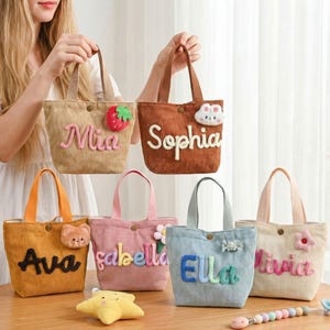 May include: A collection of corduroy tote bags in various colors, each personalized with a name in a playful font. Adorned with decorative appliqués like a strawberry, flower, and bear, these bags feature sturdy handles and a button closure.