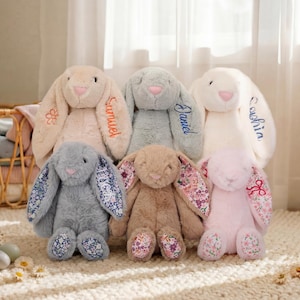 May include: Six plush bunny rabbits in various pastel colors, including pink, beige, and gray. Each bunny has long ears, embroidered details, and some feature floral patterns. Names are embroidered on some of the bunnies.