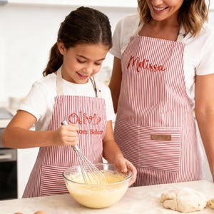 May include: Two aprons with red and white stripes. One apron has the name "Olivia" embroidered on it, and the other has the name "Melissa". A child is whisking ingredients in a glass bowl, while an adult looks on. Eggs and dough are on the counter.