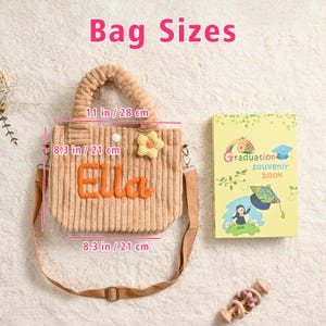 May include: A tan corduroy tote bag with the name "Ella" in orange, a yellow flower, and a detachable shoulder strap. The bag measures 11 inches wide and 8.3 inches high. A graduation souvenir book and a wooden toy are also in the image.