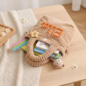 May include: A tan corduroy tote bag with the name "Ella" in orange letters. The bag is filled with books and a small stuffed bear. A ruler and a flower-shaped embellishment are visible. The bag sits on a light wooden table.