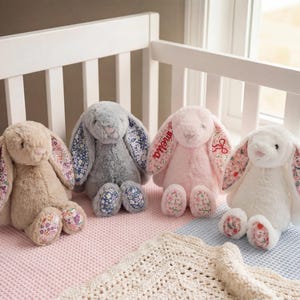 May include: Four soft toy bunny rabbits in beige, grey, pink, and white, with floral patterned ears and feet. They are arranged on a pink and blue blanket, with a white cot in the background.