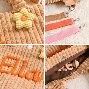 May include: A tan corduroy baby bag with a flower applique and the name "Ella" embroidered in orange. The bag has a brown interior and comes with three detachable straps in brown, orange, and pink.