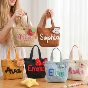 Könnte beinhalten: Eine Sammlung von Cord-Tragetaschen in verschiedenen Farben, jede mit einem Namen in einer verspielten Schriftart personalisiert. Verziert mit dekorativen Applikationen wie einer Erdbeere, einem Bären und einer Blume. Die Taschen haben stabile Griffe und einen Knopfverschluss.