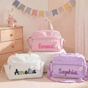 May include: Three baby bags in pastel colors: pink, white, and lavender. Each bag has a name embroidered on the front: "Emma", "Amelia", and "Sophia". The bags have a flower or cloud embellishment. A stuffed bunny sits on a wooden dresser.