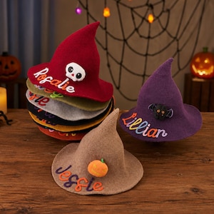 May include: A collection of knitted witch hats in various colors including red, purple, and tan. Each hat features a personalized name embroidered in cursive script and a decorative felt applique such as a skull, a bat, or a pumpkin.