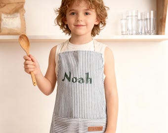 Puede incluir: Un delantal a rayas azules y blancas para niños, bordado con el nombre "Noah" en verde. Tiene un bolsillo delantero y una pequeña etiqueta de cuero. El niño sostiene una cuchara de madera.