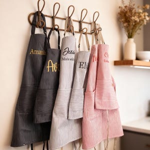 Könnte beinhalten: Eine Auswahl an gestreiften Schürzen in Schwarz, Grau und Rot mit gestickten Namen, die an einem dekorativen Gestell hängen. Die Schürzen haben Taschen und lange Bindebänder. Die Namen sind in Gold und Schwarz gestickt.