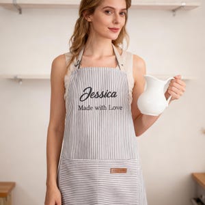 May include: A grey and white vertical striped apron featuring the embroidered name Jessica and the text Made with Love in black thread. The apron includes a large front pocket with a small brown rectangular leather-like patch on the right side.